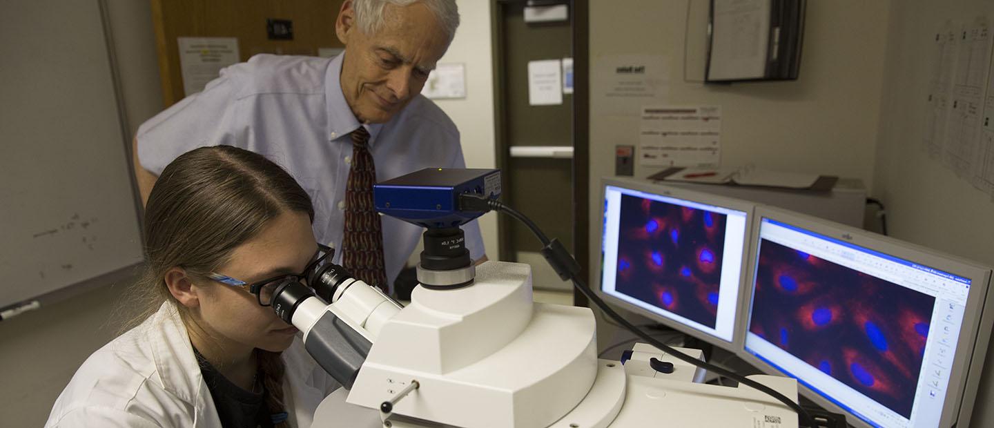 professor standing over a student looking in a microscope with two computer display screens