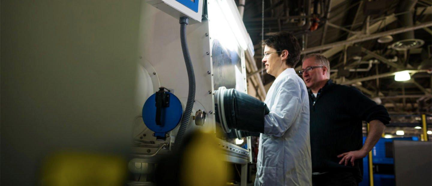 A woman in a white lab coat working at a large machine while a man watches over her shoulder.